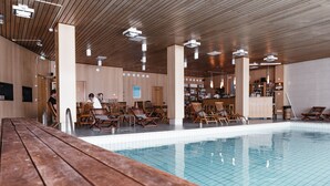Un área de piscina con mesas y sillas de jardín de madera, un bar con estantes llenos de botellas, y un techo de azulejos.