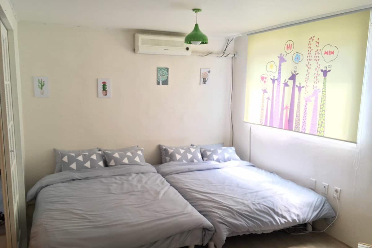 Ein Schlafzimmer mit zwei Betten, einer grünen Pendelleuchte und einem Fenster mit Wandgemälde.