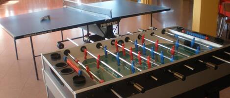 Una mesa de ping pong y una mesa de fútbolín en una sala con sillas y una escalera al fondo.
