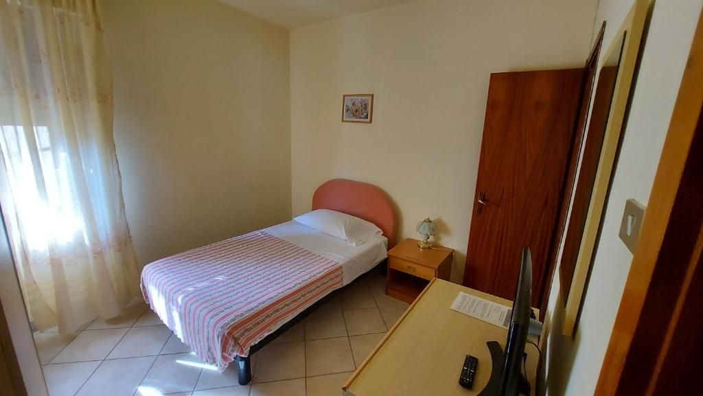 A single bed with a striped blanket, a bedside table with a lamp, a wooden wardrobe, and a desk with a computer monitor.
