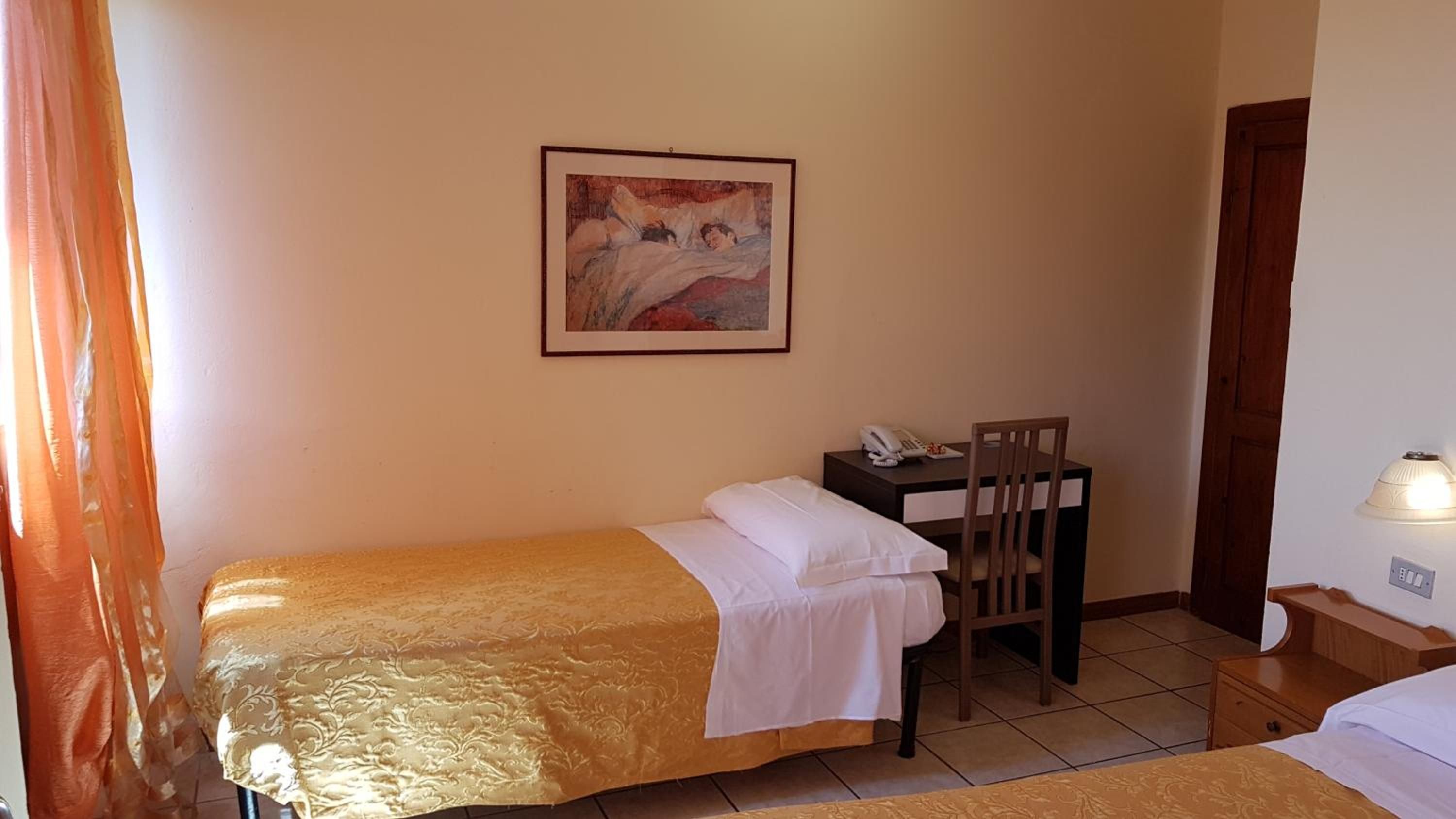 Una cama individual con cubrecolor dorado, un escritorio con silla, un cuadro en la pared y una lámpara en la mesita de noche.
