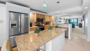 A modern kitchen with a stainless steel refrigerator, granite countertop, and wooden cabinets. There are two potted plants on the island and pendant lights above.
