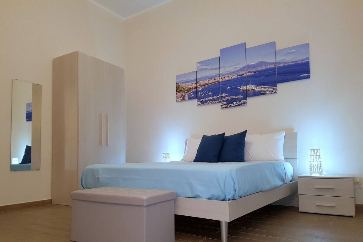 A modern bedroom with a bed, a nightstand, a bench, and a wall-mounted artwork depicting a coastal scene.