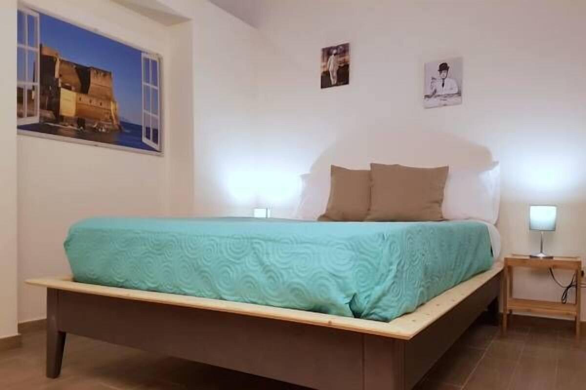 A bedroom with a bed, a nightstand, a wall-mounted lamp, and a poster of a shipwreck.