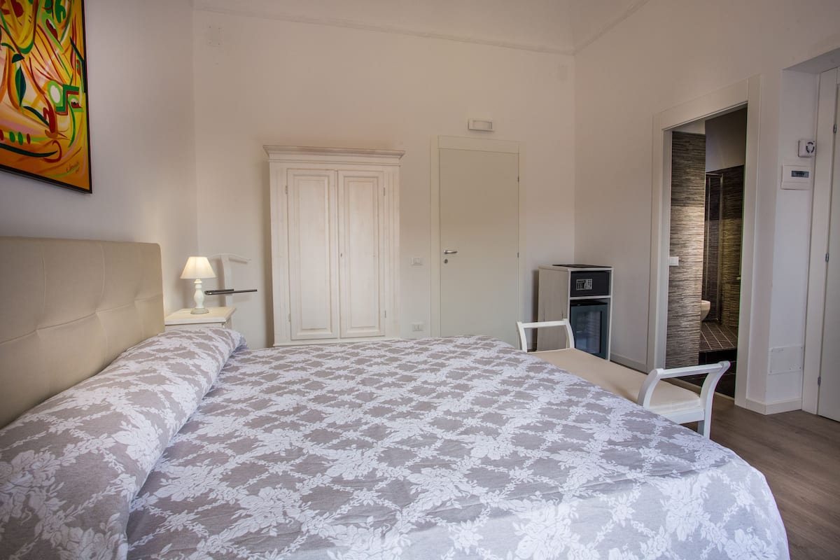 Un dormitorio con cama, mesita de noche, silla, armario y baño visible a través de una puerta abierta.