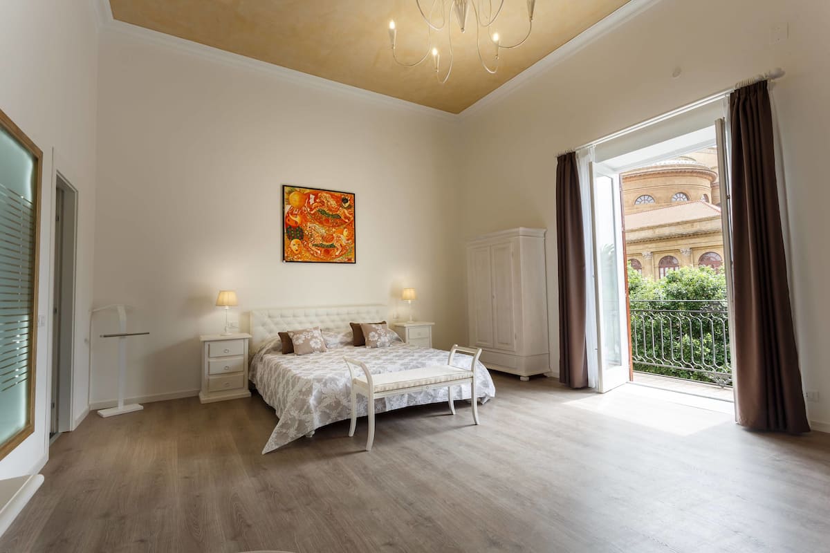 Un dormitorio amplio con una cama grande, una mesita de noche, una silla, un cuadro y vistas a un edificio a través de una puerta abierta.