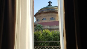 Vista per una finestra amb cortines, que mostra un edifici amb una cúpula i un balcó amb barana metàl·lica.