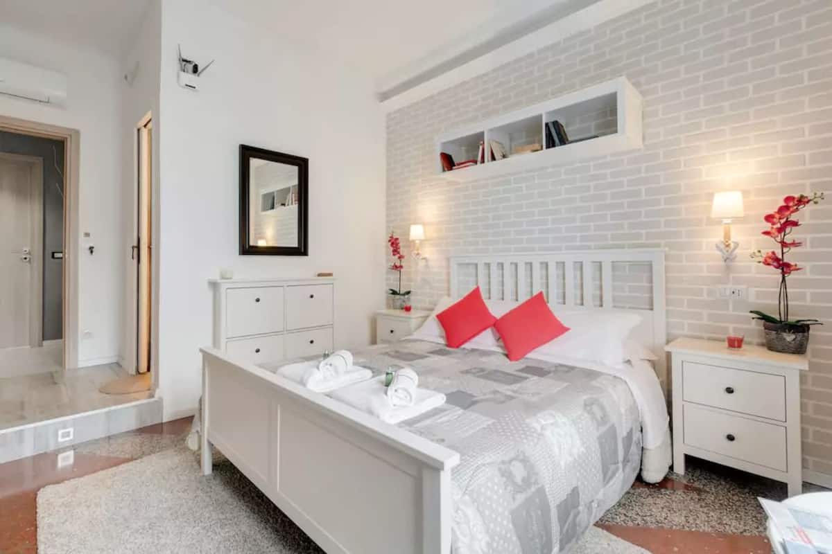 Ein Schlafzimmer mit weißer Ziegelwand, einem Bett mit weißer Bettwäsche und roten Kissen, einem Nachttisch mit Lampe und einem kleinen Regal über dem Bett.
