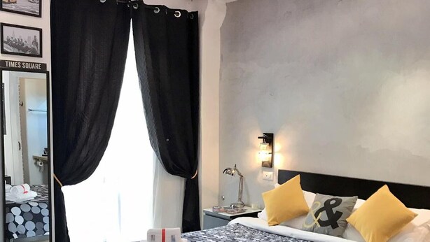 A modern hotel room with a large bed, black and white patterned bedding, a nightstand with a lamp, and a mirror with a Times Square sign. - Moonlight Inn Guest House (Rome)