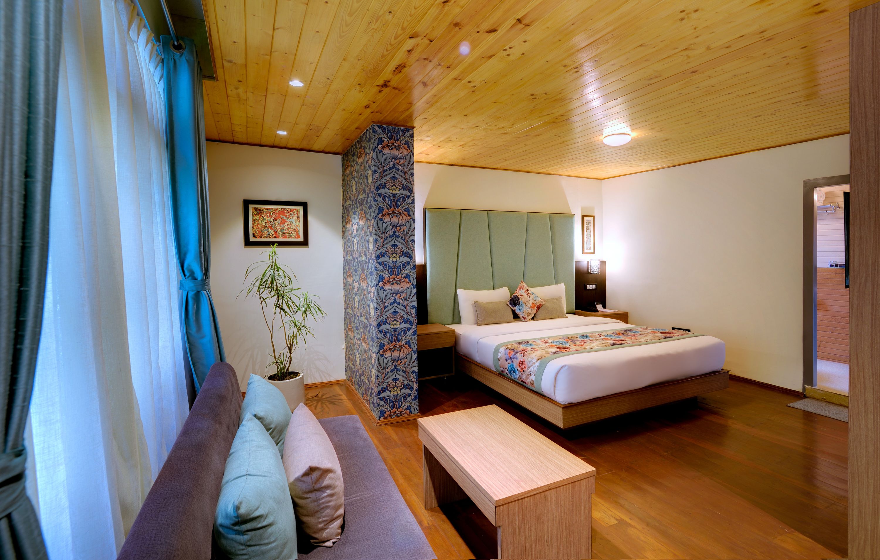 A bedroom with a wooden ceiling, a bed with a floral bedspread, a bench, a nightstand, and a potted plant.