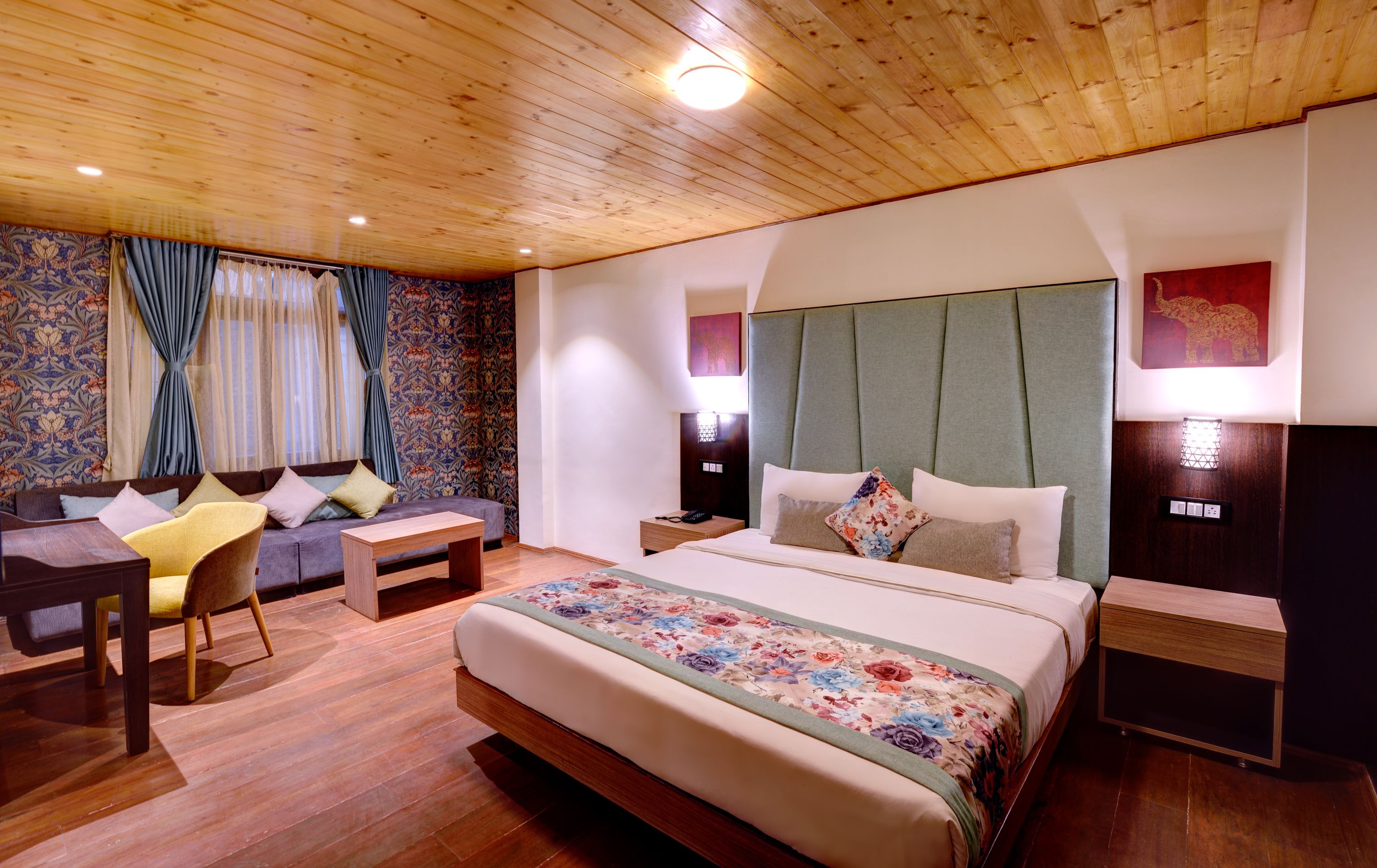 A hotel room with a wooden ceiling, a bed with floral bedding, a sofa with cushions, a small table, and a nightstand with a lamp.