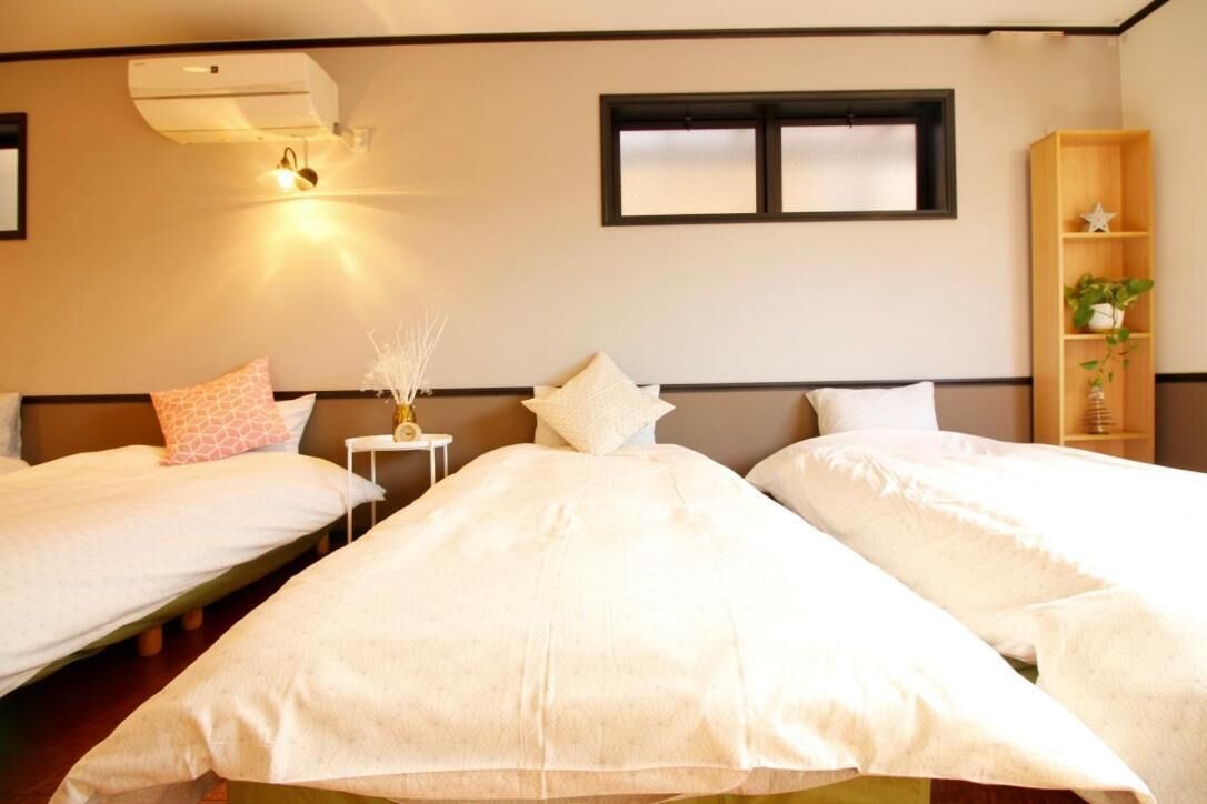 A hotel room with two single beds, a small bedside table with a lamp, a window, and a wooden shelf with a plant.