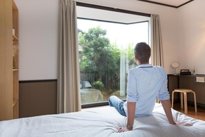 A man sitting on a bed in a room with a large window, a wooden shelf, and a small stool. - Guesthouse Meguri Inari (Kyoto)