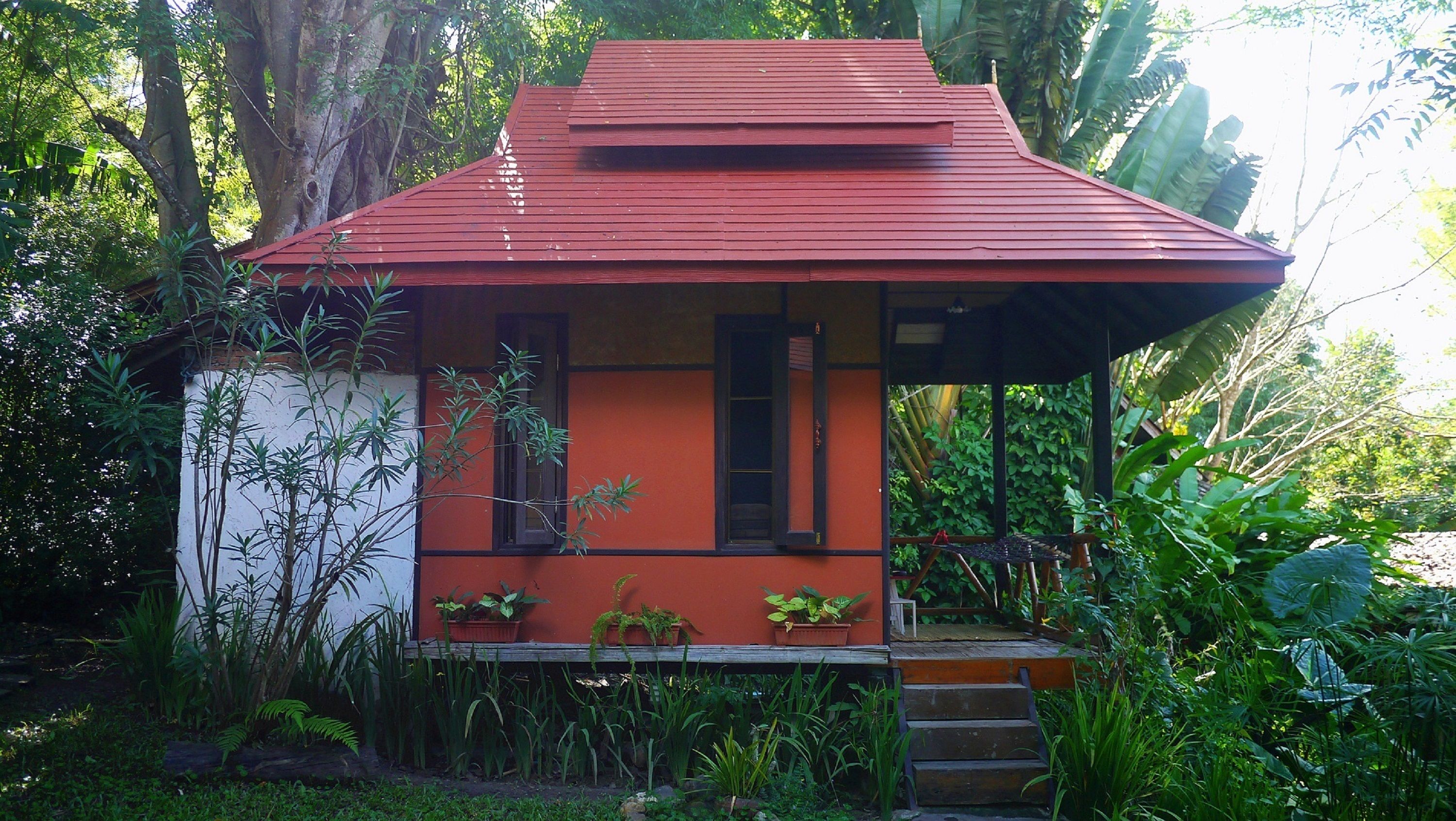 A small, single-story house with a red roof and white walls, surrounded by lush greenery.