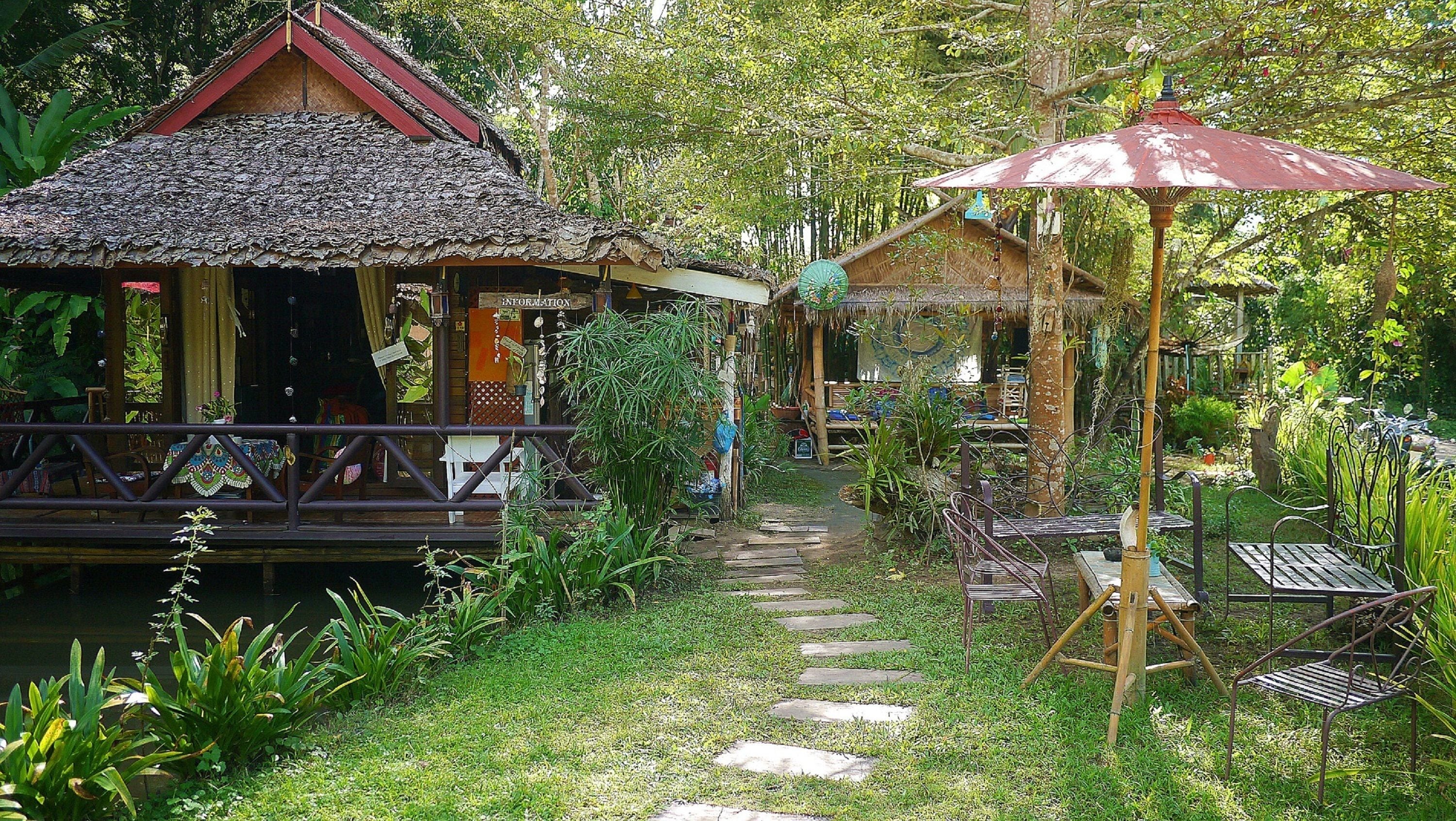A rustic wooden structure with a thatched roof, a covered seating area with a metal umbrella, and a stone pathway leading through a lush garden.