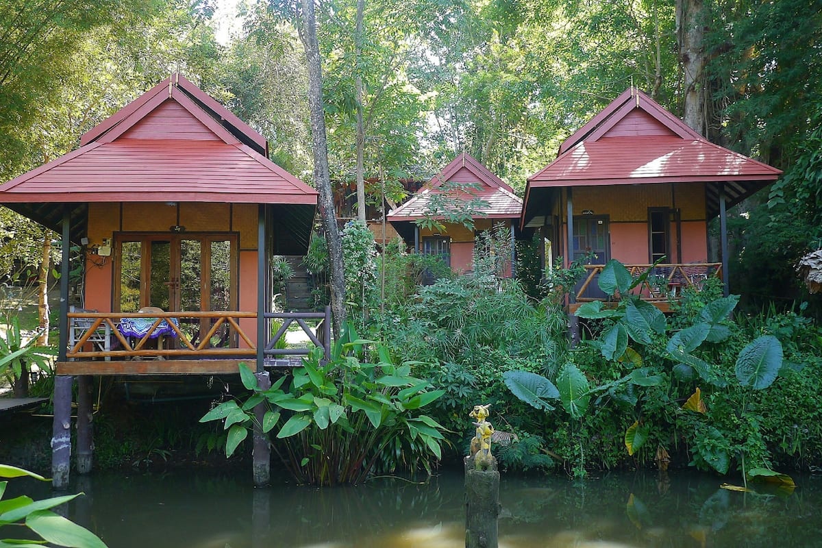 Three wooden cabins with red roofs, surrounded by lush greenery and a body of water.