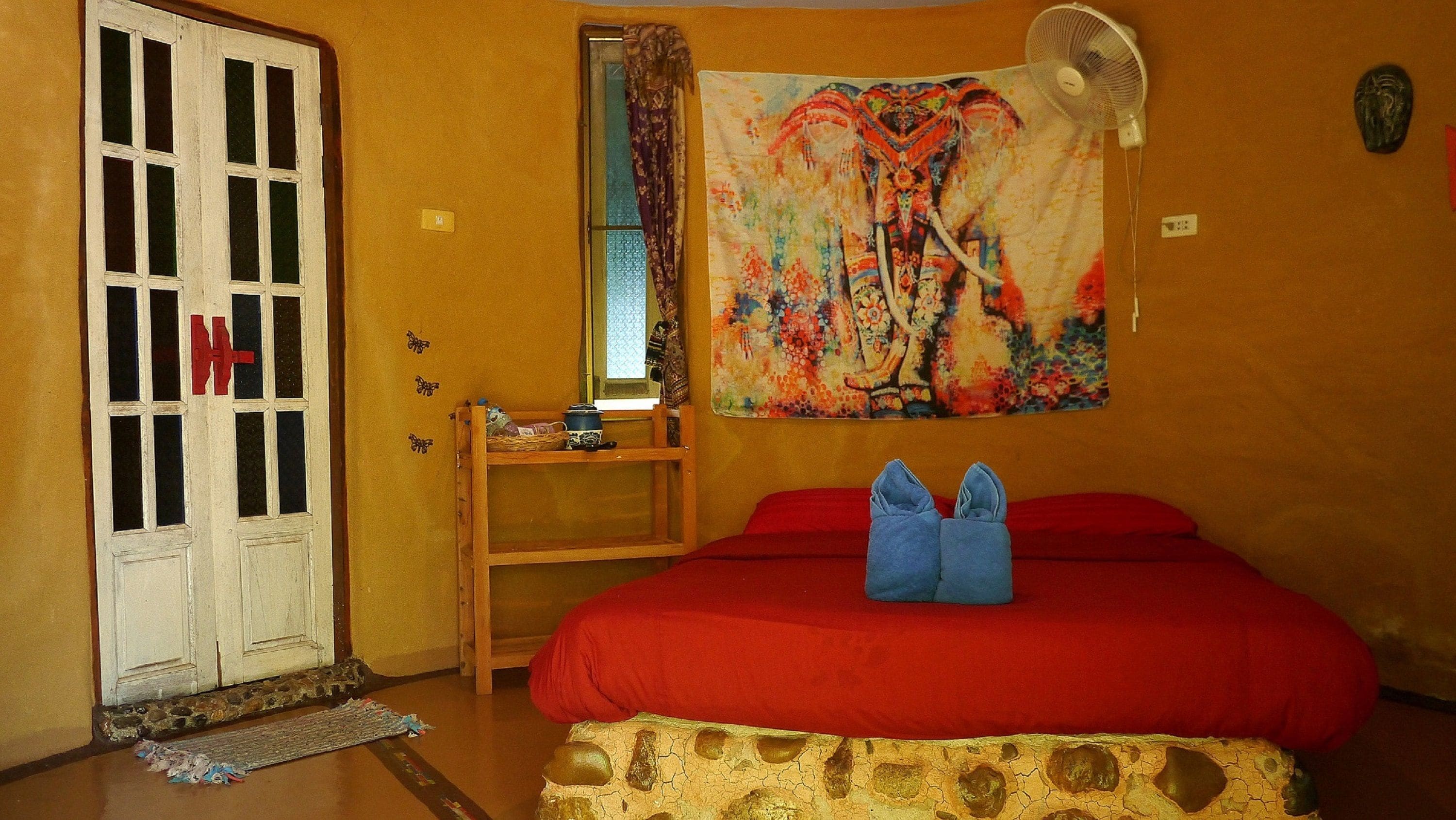 A room with a bed, a fan, a tapestry, a door, and a small table with items on it.