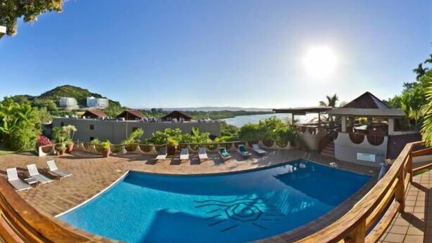 A pool with a wooden deck and lounge chairs, surrounded by buildings and a view of the water. - Cliffside View Hotel (Meyungs)