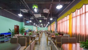 A spacious indoor area with multiple tables and chairs arranged in rows, a few potted plants, and a large window with curtains. - Guilin Manhattan Hotel North Station (Guilin)