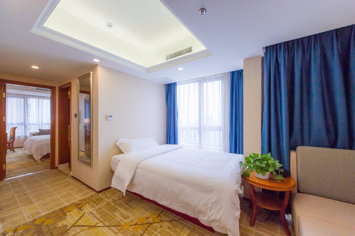 Ein Hotelzimmer mit einem großen Bett, einem kleinen Sofa, einem Beistelltisch mit Pflanze, einem Spiegel und Blick ins Freie durch ein Fenster mit blauen Vorhängen.