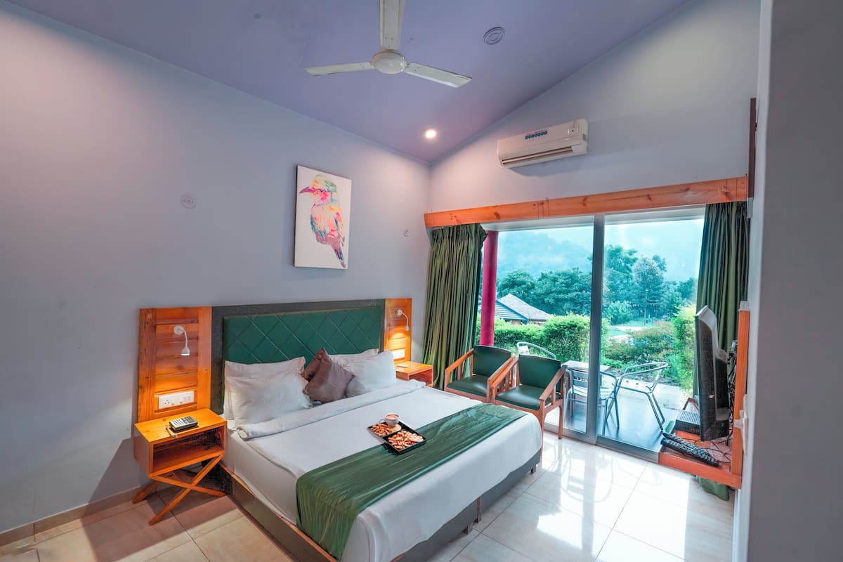A hotel room with a bed, bedside tables, a desk with a chair, a window with curtains, and a view of greenery outside.