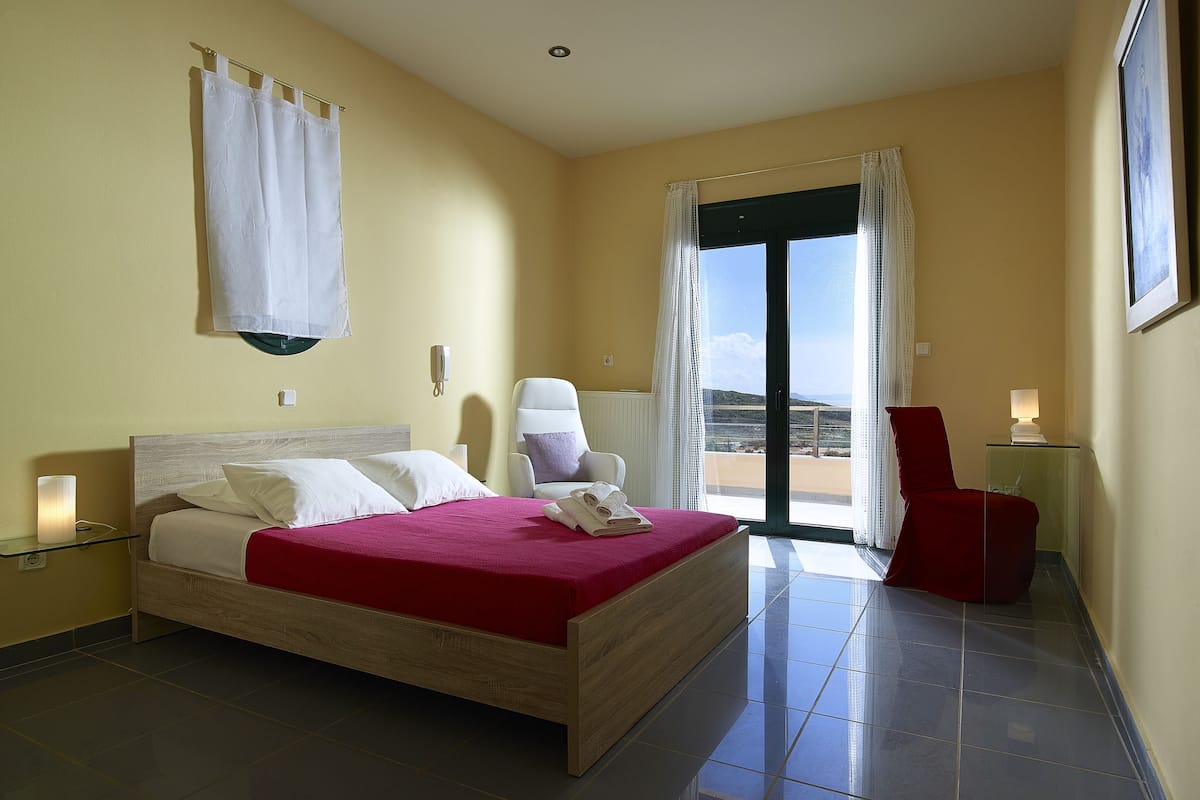 A bedroom with a bed, a chair, a small table, and a view of the outdoors.