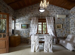 A rustic room with stone walls, a wooden ceiling, a chandelier, a bed, a table with a chair, and two windows with curtains.