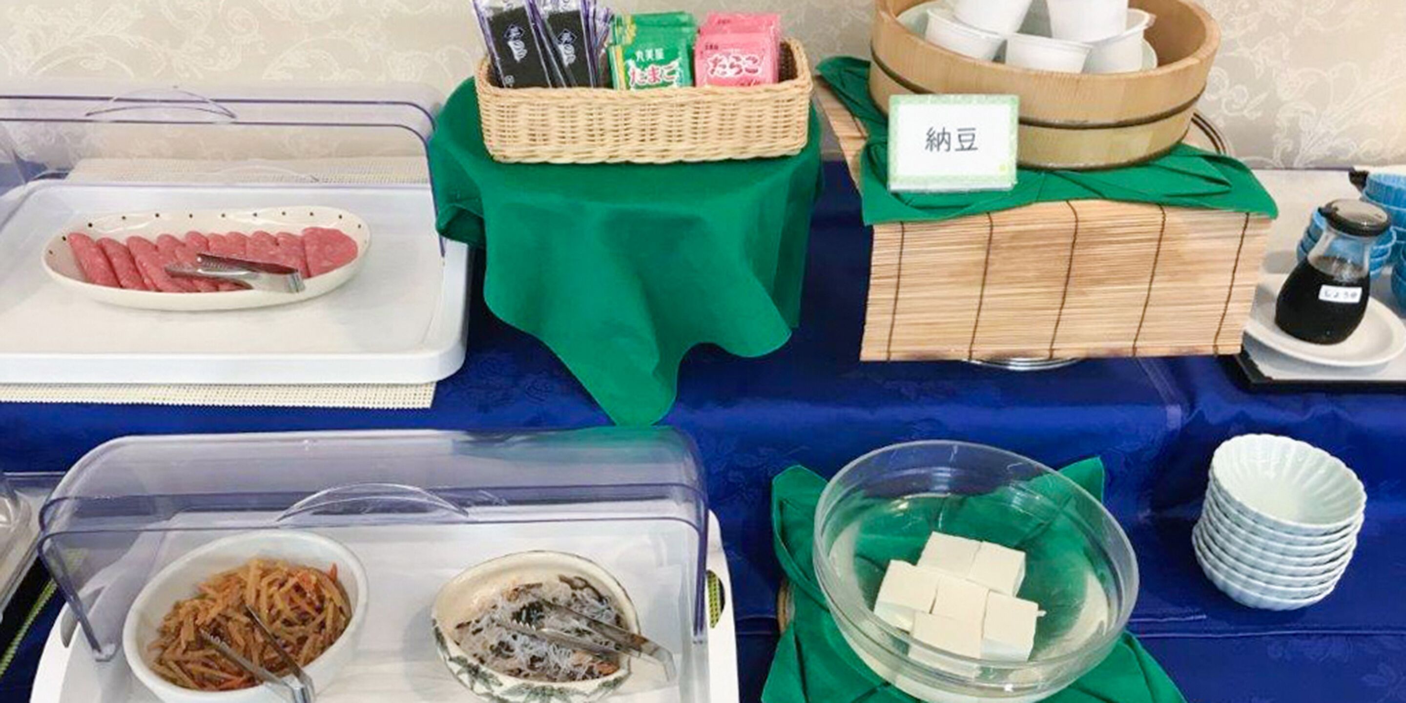 A buffet table with various food items and utensils, including a plate of sliced meat, a bowl of salad, and a container of tofu.
