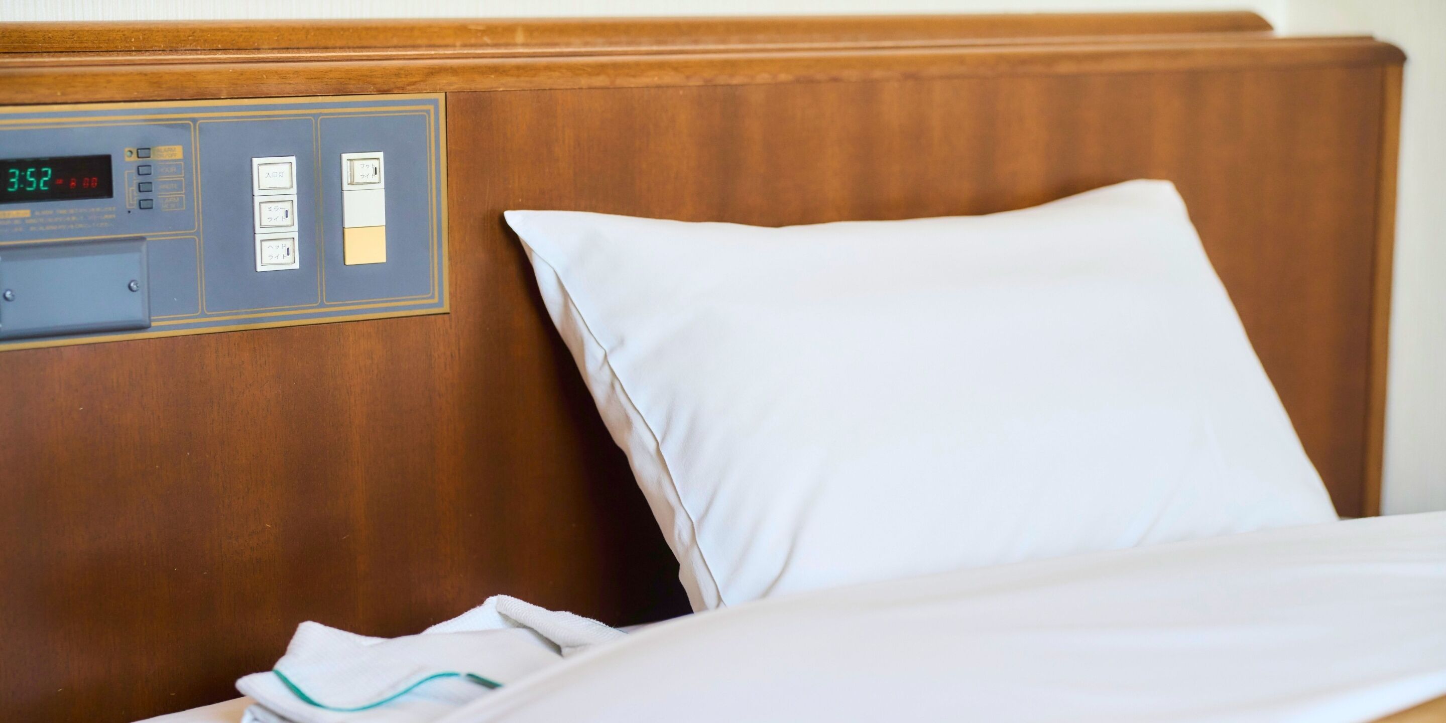 Una habitación de hotel con cabecera de madera, un reloj digital y una almohada blanca.