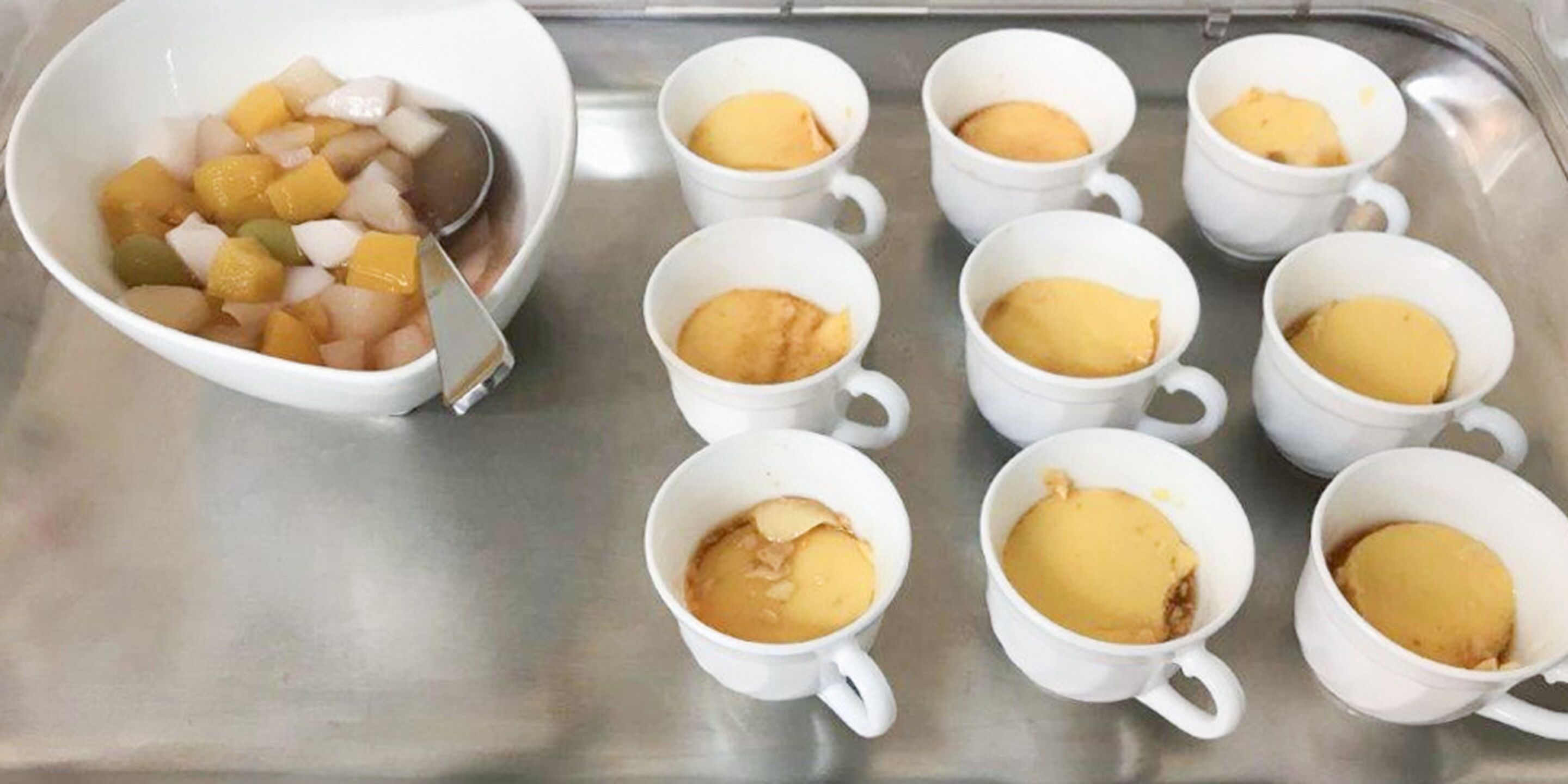 Eine Tablett mit verschiedenen weißen Tassen, gefüllt mit gelbcremigen Desserts, und einer Schüssel mit gewürfelten Früchten und einem Löffel.