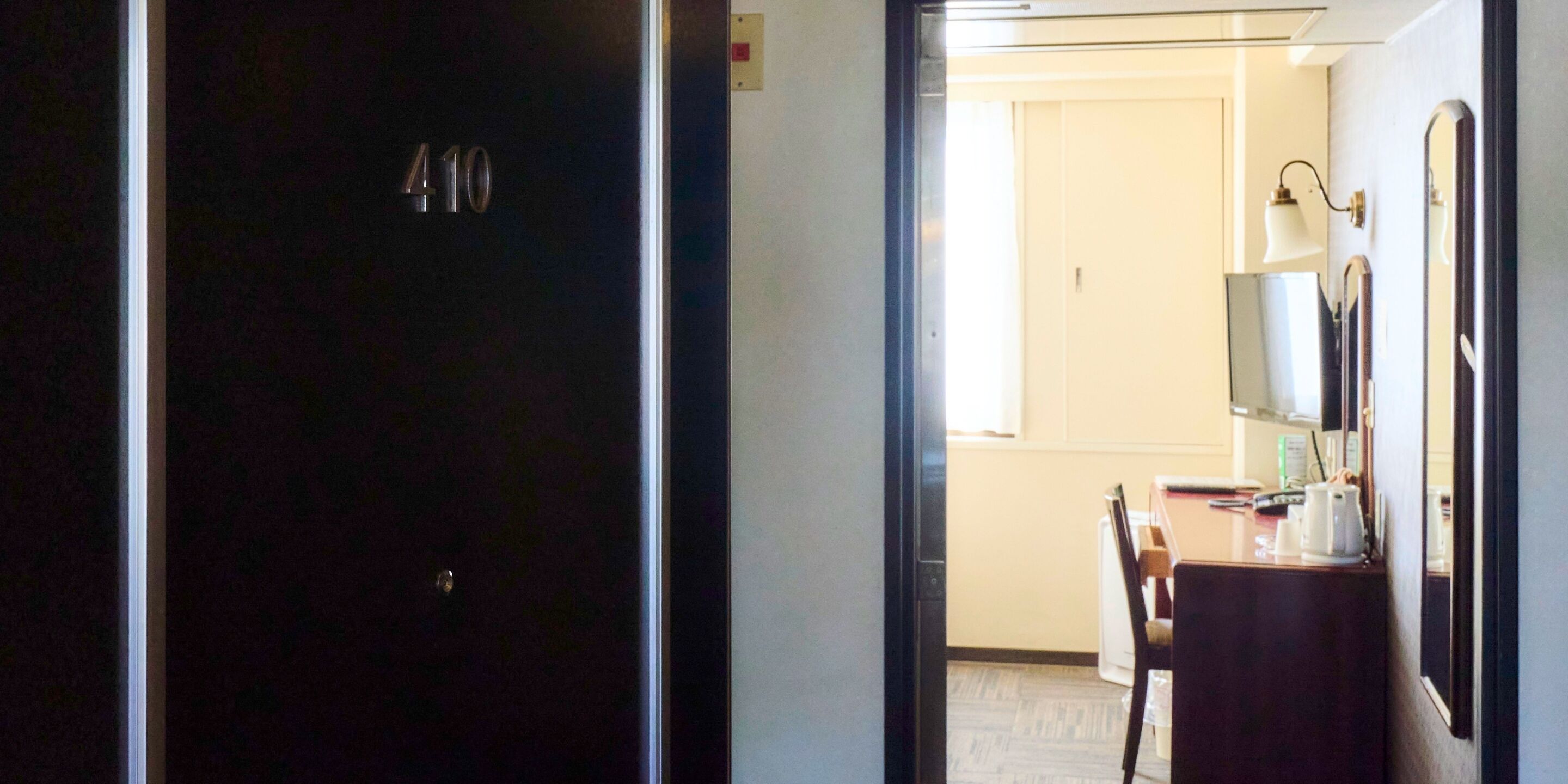 A hotel room with a dark door, a small table with a chair, a desk with a lamp, and a mirror on the wall.