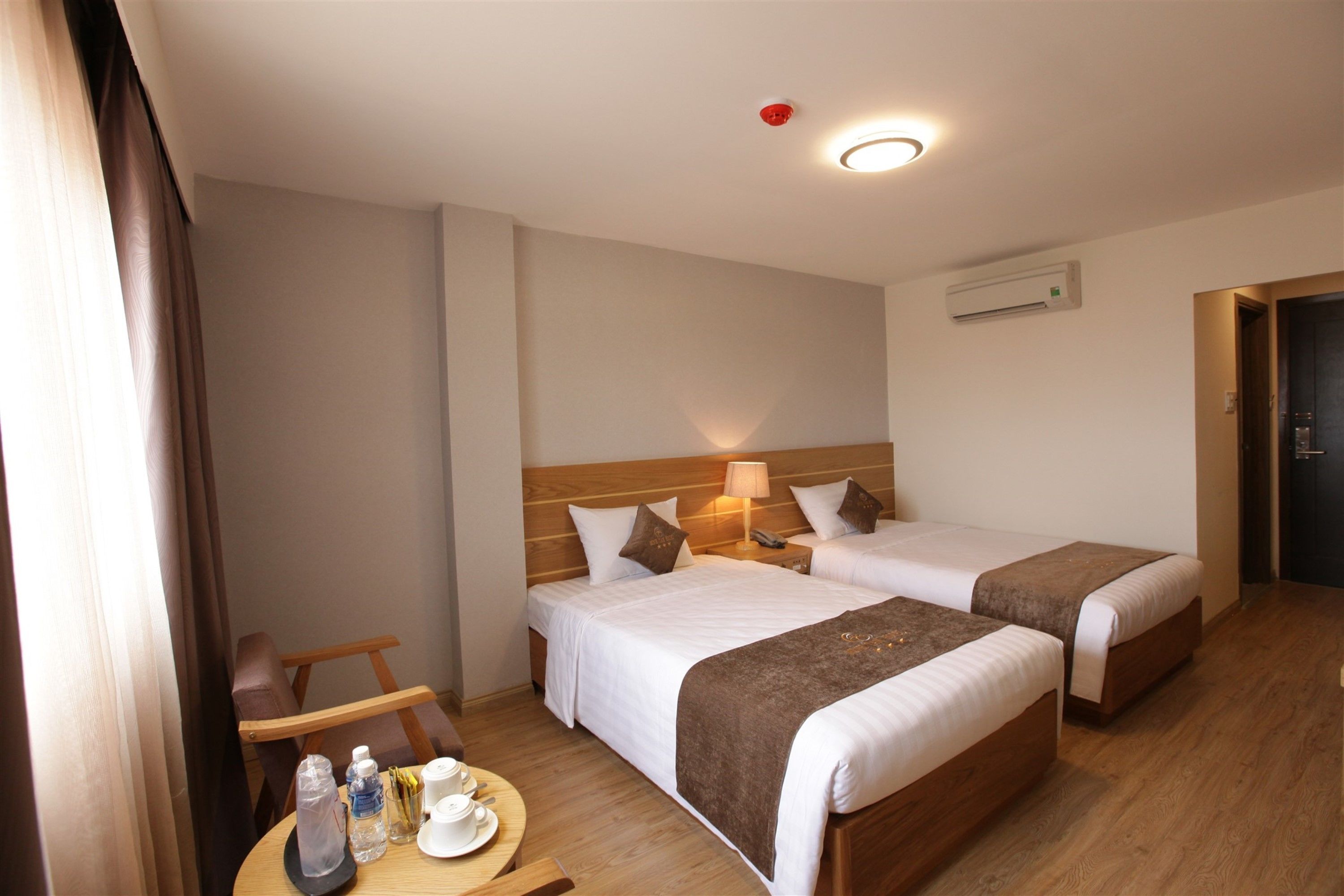 Habitación de hotel con dos camas, cabecera de madera, mesitas de noche con lámparas, una mesa redonda con bandeja y una botella de agua.