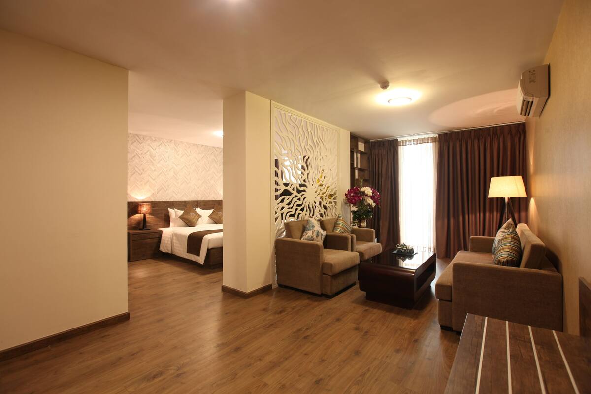 A hotel room with a wooden floor, a bed with white bedding, two sofas, a coffee table, a decorative partition, and a lamp.