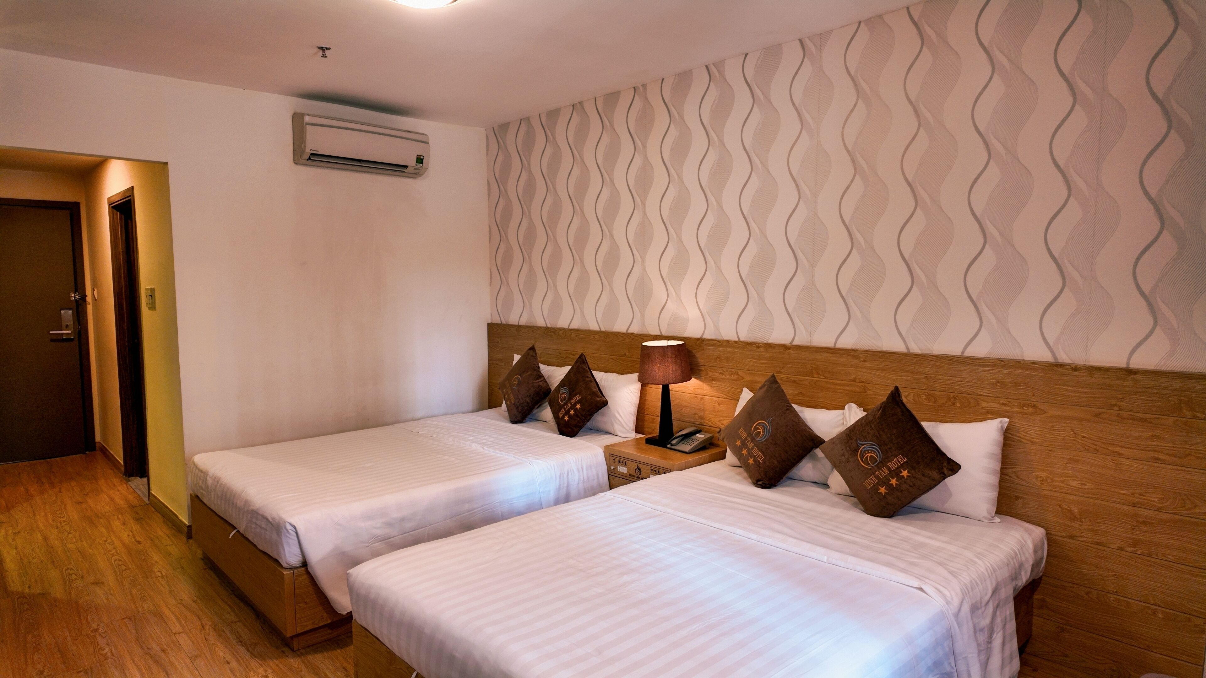 A hotel room with two beds, a wooden headboard, a nightstand with a lamp, and a patterned wall.