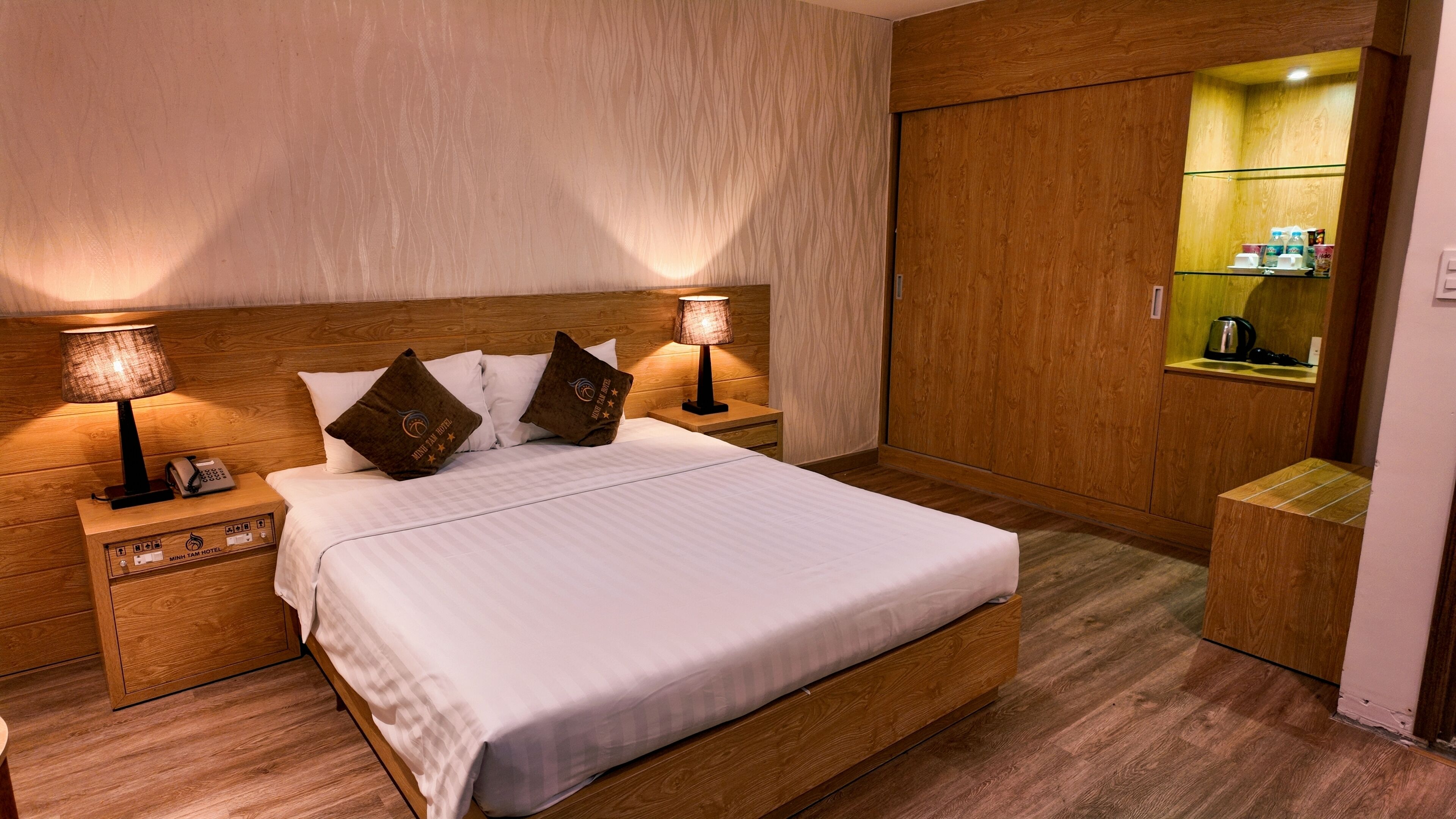 A hotel room with a wooden bed, two bedside tables with lamps, a minibar, and a wooden wardrobe.