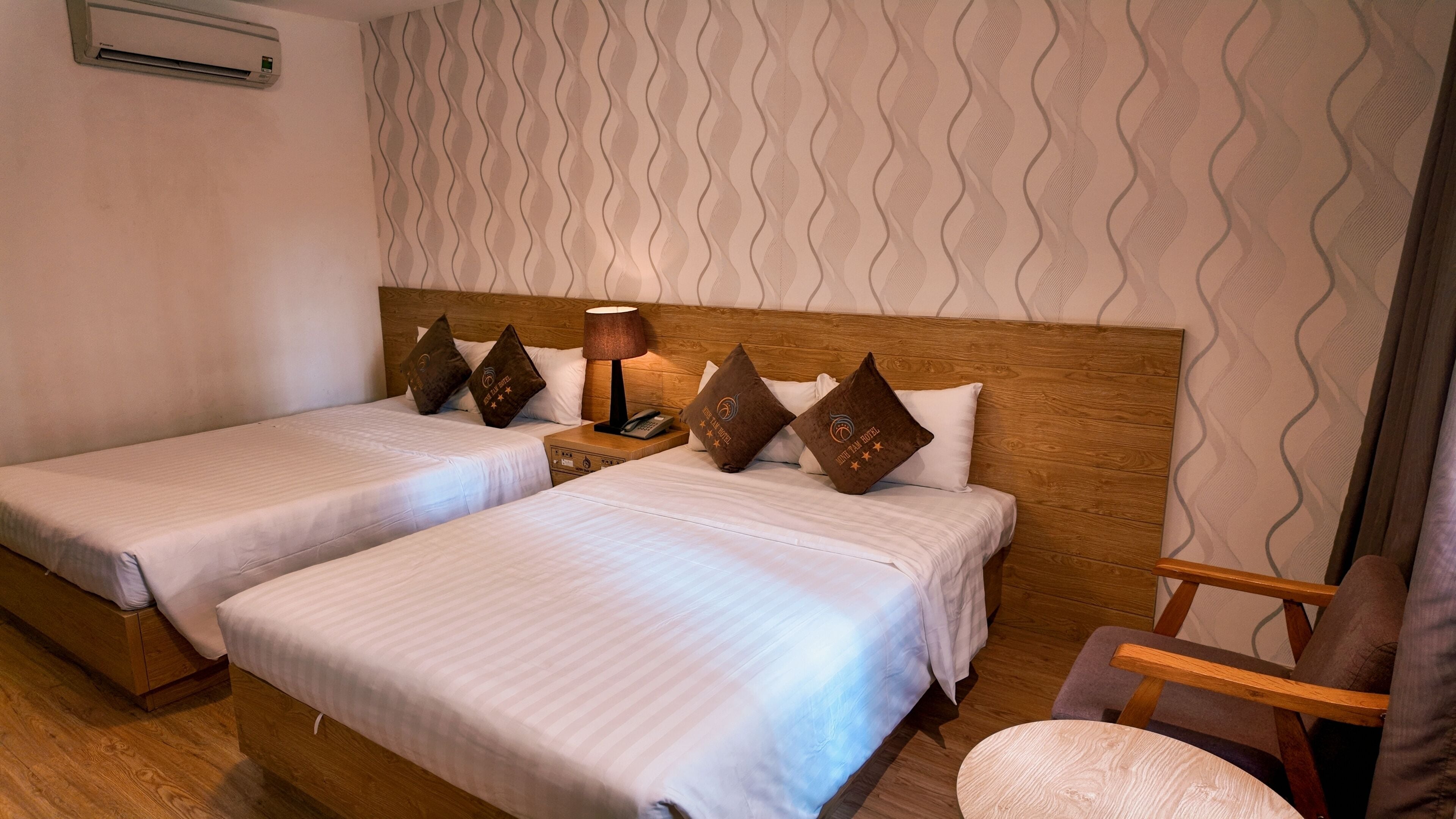 A hotel room with two beds, a wooden headboard, a nightstand with a lamp, and a chair.