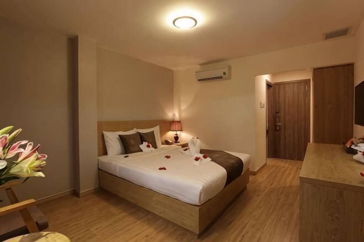 A hotel room with a bed, bedside table, lamp, and a wooden floor.