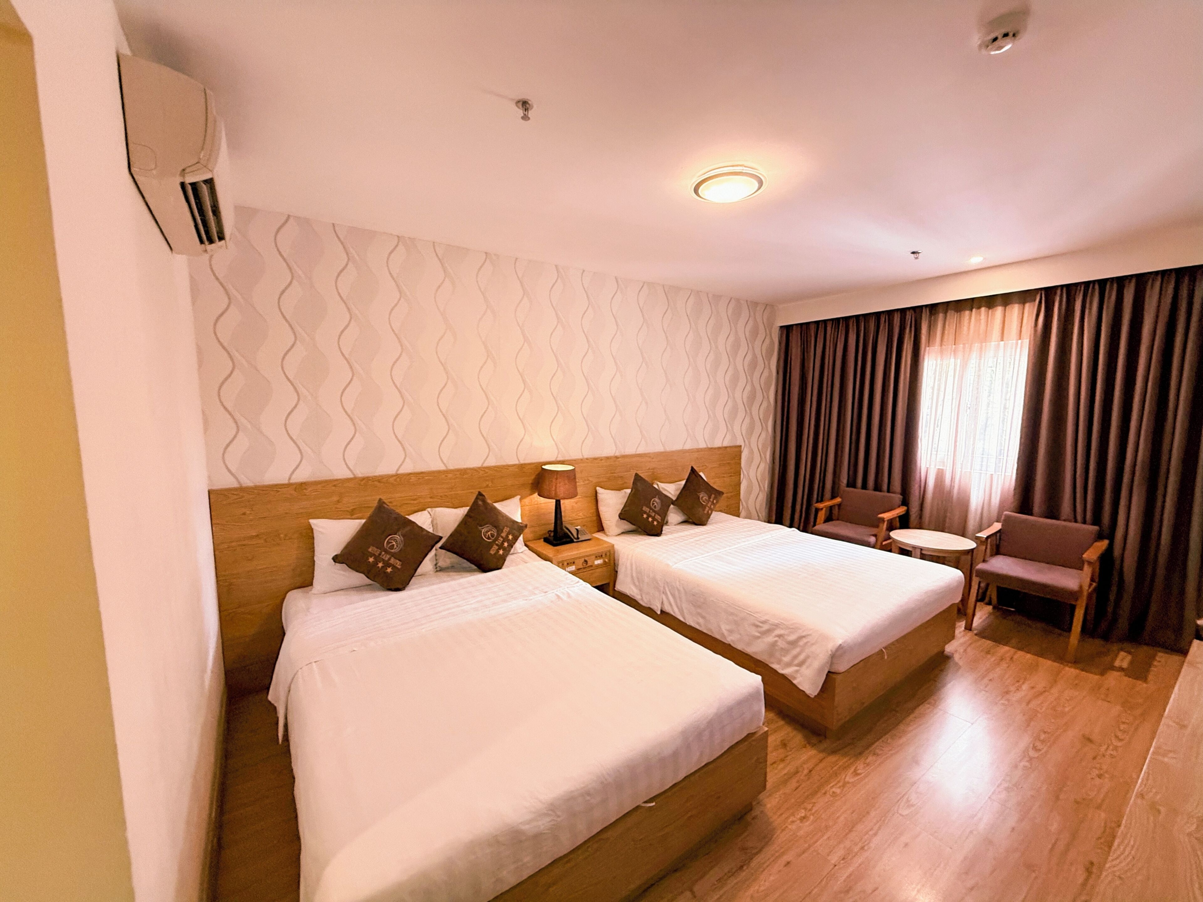 Ein Hotelzimmer mit zwei Betten, einem kleinen Tisch, einem Stuhl und einem Fenster mit Vorhängen.