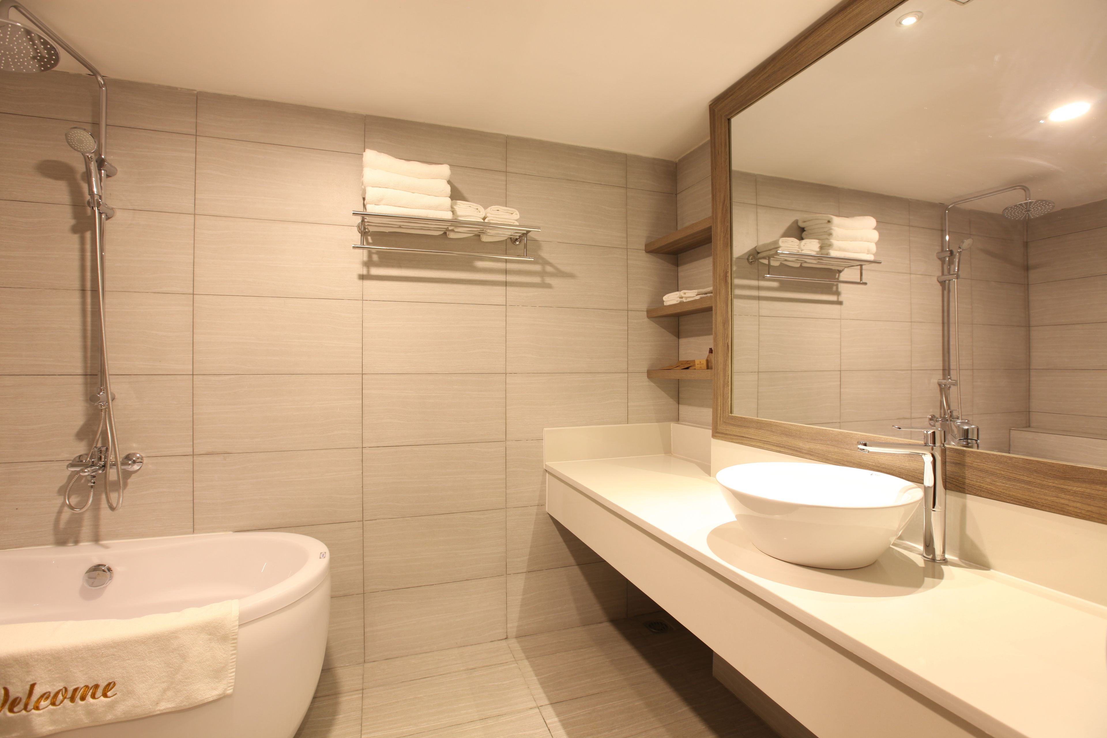 A modern bathroom with a bathtub, a large mirror, and a sink with a bowl. There are shelves with folded towels and a "Welcome" towel on the bathtub.