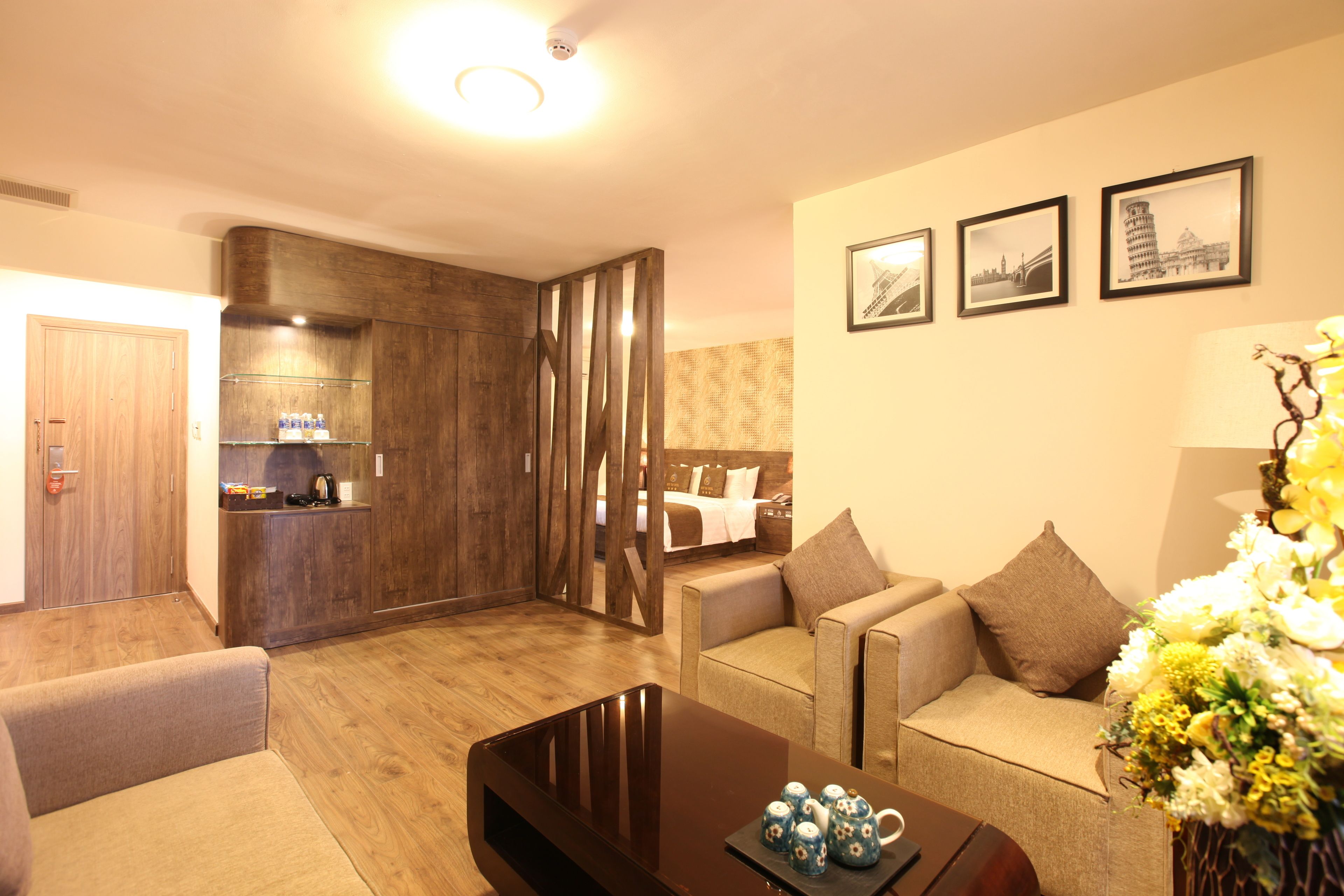 A modern living room with a sofa, a coffee table, and a dining area with a wooden cabinet. There are framed pictures on the wall and a vase with flowers on the right side.