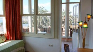 Una habitación con cama, ventana con cortinas y vista a palmeras y un edificio.