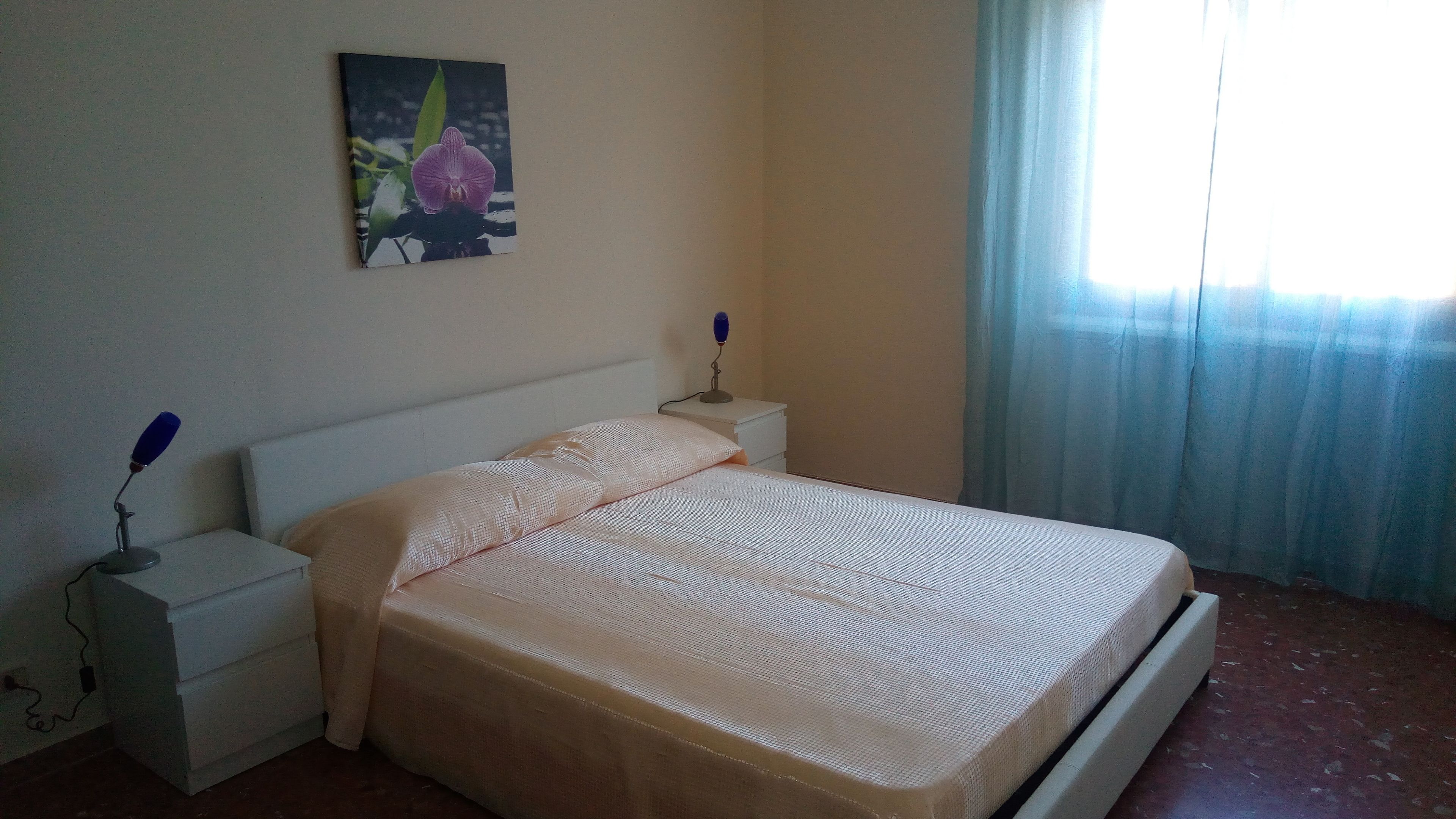Un dormitorio con una cama, dos mesitas de noche, una lámpara de noche, un cuadro en la pared y una ventana con cortinas.