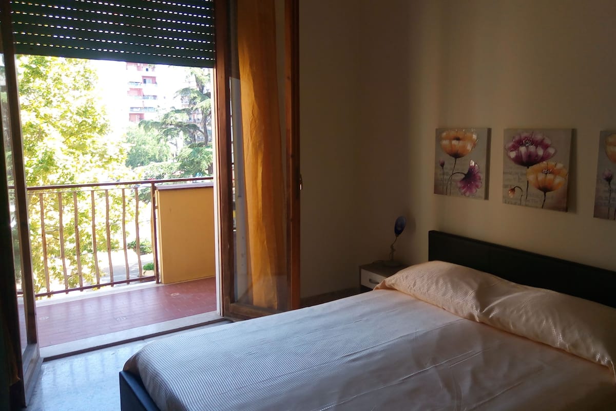 A bedroom with a bed, a nightstand, and a window with a view of a balcony and buildings.