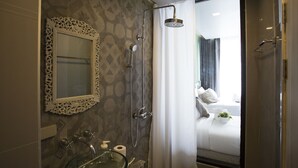 A bathroom with a glass sink, a mirror with an ornate frame, a shower with a handheld showerhead, and a view into a bedroom with a bed and a window. - Heyha Hotel (Bangkok)