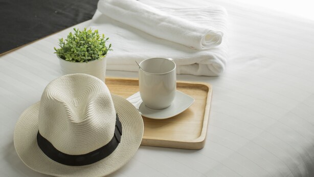 A neatly made bed with a white and black bedspread, a small potted plant, a folded white towel, a white cup, and a white hat on a tray. - Heyha Hotel (Bangkok)