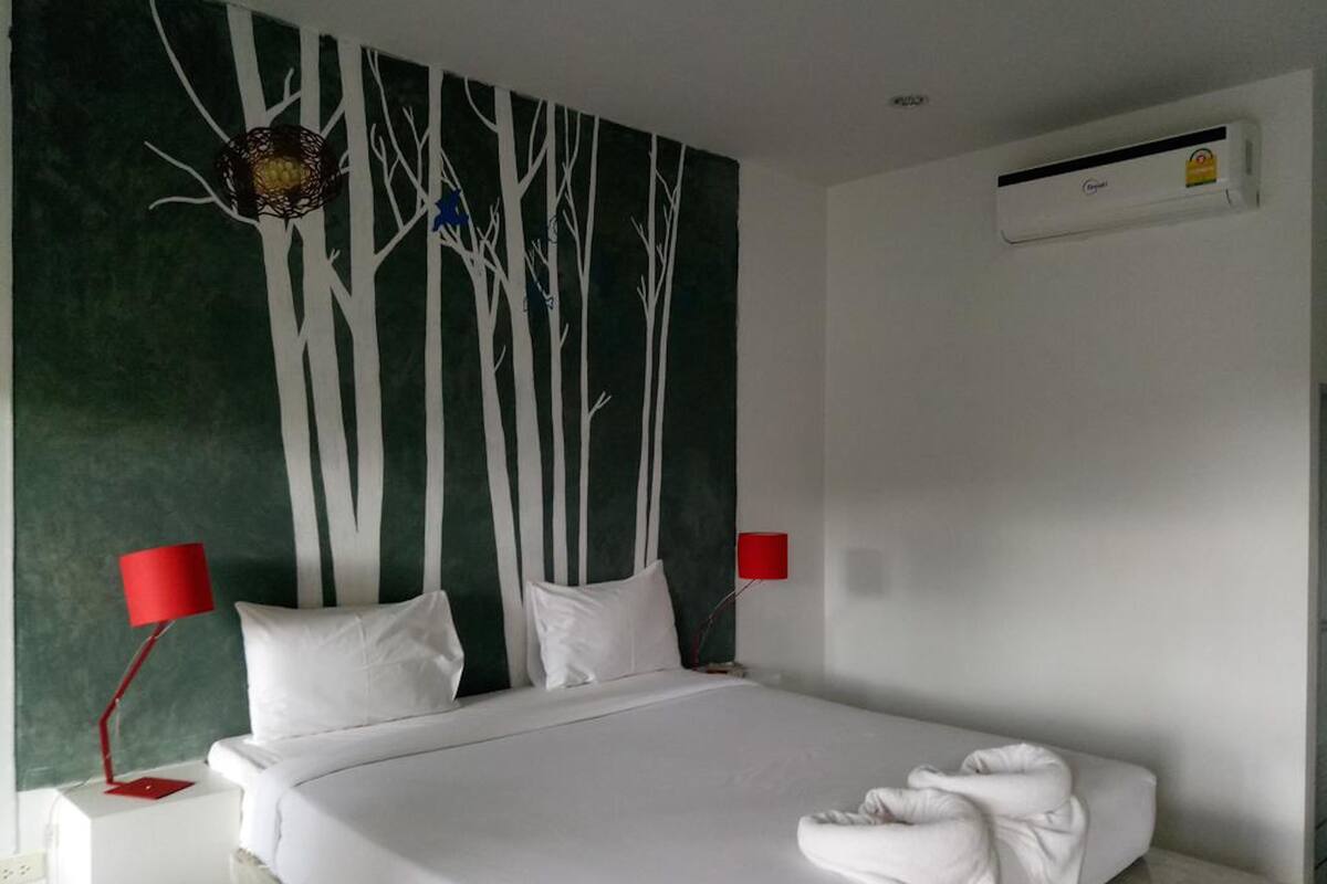 Habitación de hotel con cama, dos lámparas de noche rojas, aire acondicionado de pared y un cuadro grande con árboles.