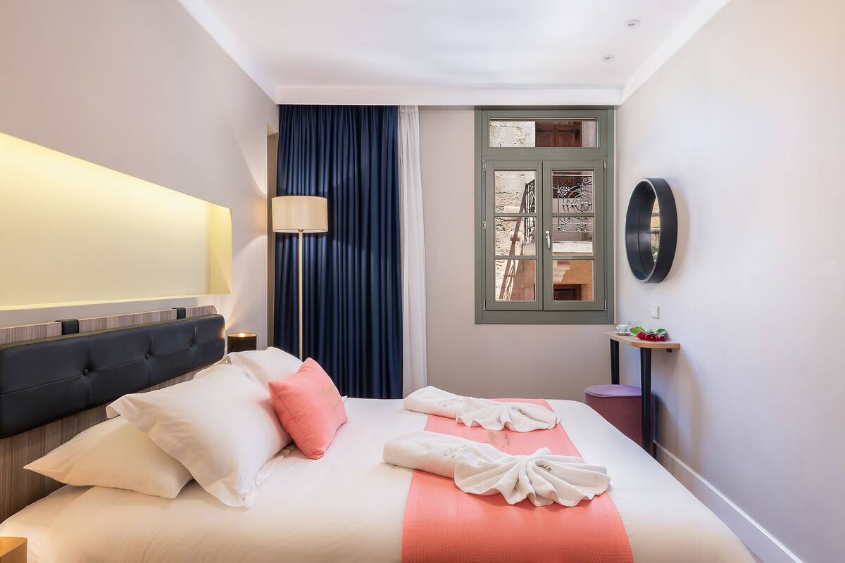 Une chambre d’hôtel avec un lit, une table de chevet, une lampe, une fenêtre avec des rideaux et une étagère fixée au mur, sur laquelle sont disposés des fruits.