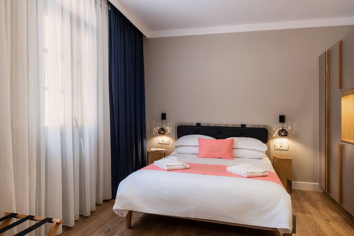 Un lit bien fait, avec du linge de lit blanc et un coussin rose, deux tables de chevet avec des lampes, un plancher en bois et une grande fenêtre ornée de rideaux légers.