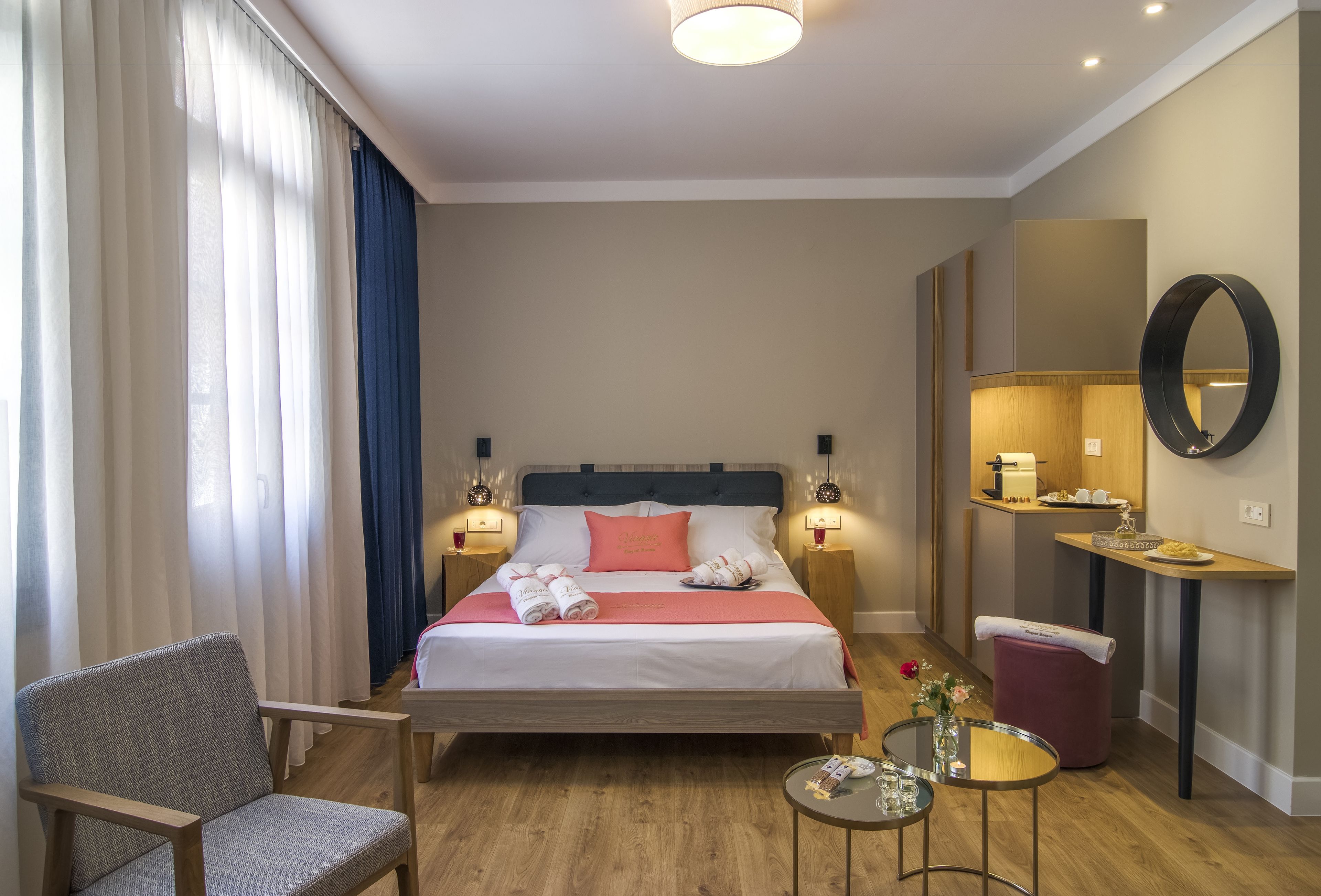 A hotel room with a bed, a chair, a small table, a mirror, and a bedside table with a lamp.
