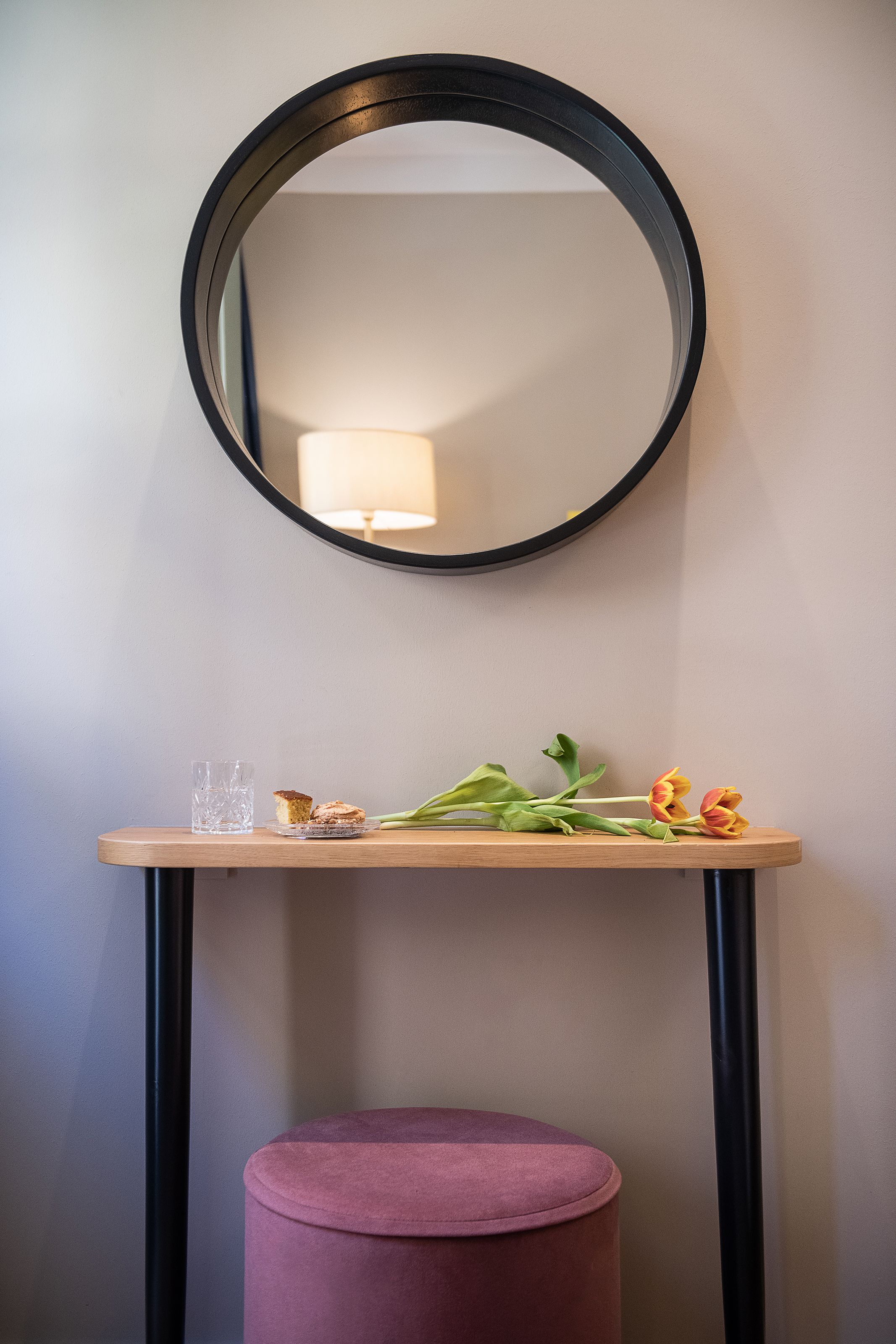 A round mirror on a wall, a small table with a glass and food, and a pink ottoman.